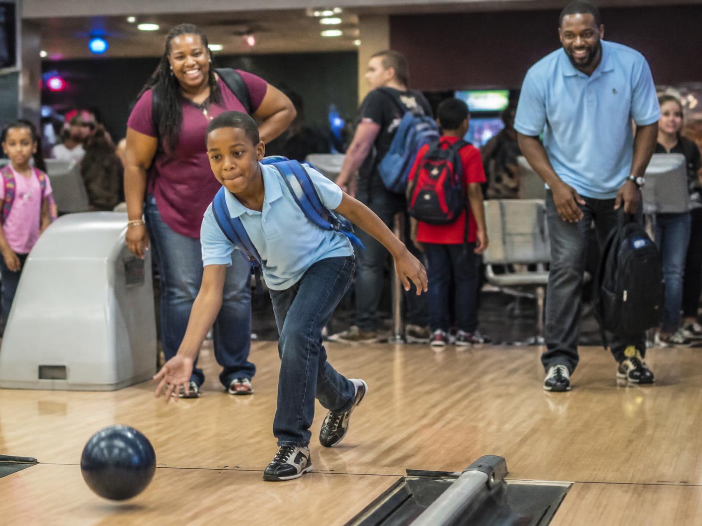 A child wearing a new backpack bowls while families and volunteers cheer.